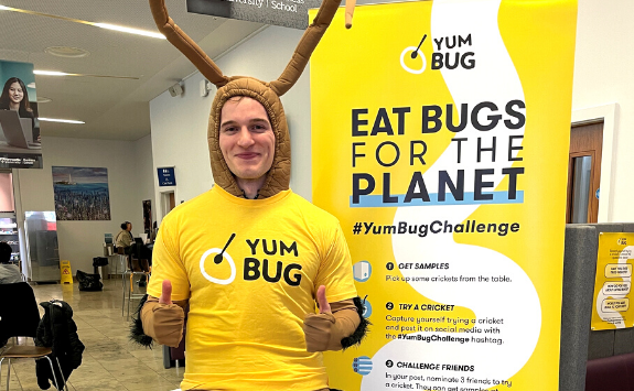 Yum Bug cofounder, Aaron, dressed up in an insect costume, wearing a Yum Bug shirt, next to a promotion banner about Yum Bug. He is in the NUBS Cafe.